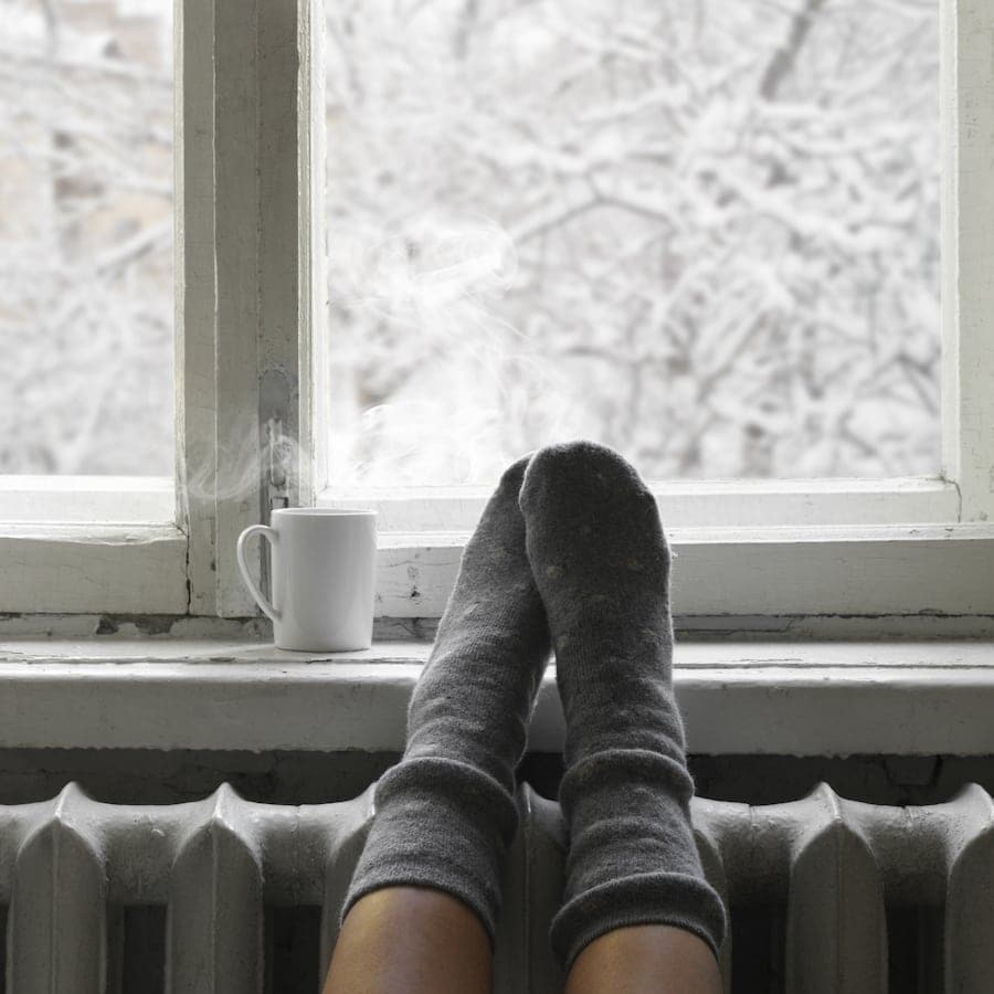 Cozy winter still life: woman feet in warm woolen socks and mug of hot beverage on old windowsill against snow landscape from outside. (Cozy winter still life: woman feet in warm woolen socks and mug of hot beverage on old windowsill against snow land.